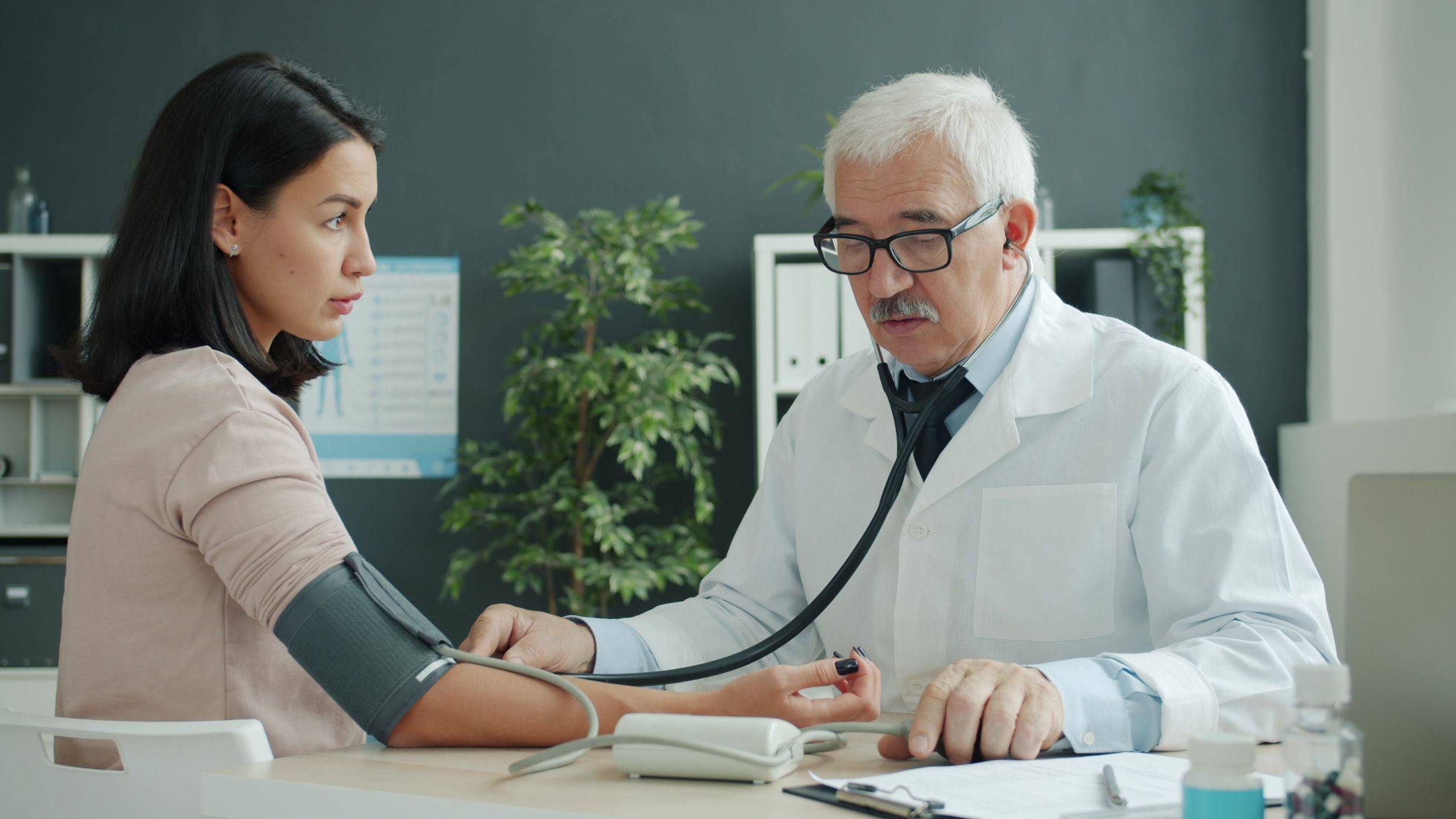 A clinician takes a patient’s blood pressure across a desk in a bright exam room.